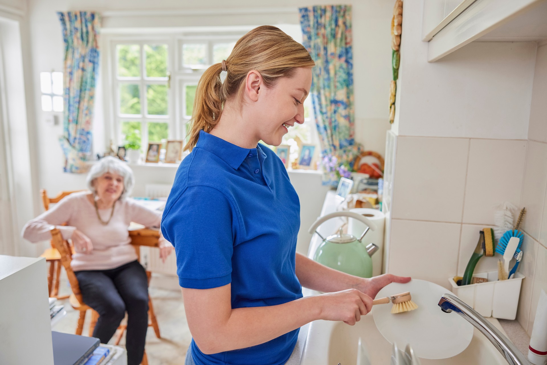 Female Home Help Cleaning House Doing Washing Up In Kitchen Whilst Chatting With Senior Woman